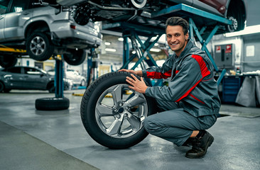 Plakat Handsome mechanic in uniform is working in auto service. Car repair and maintenance. Holding car wheel.