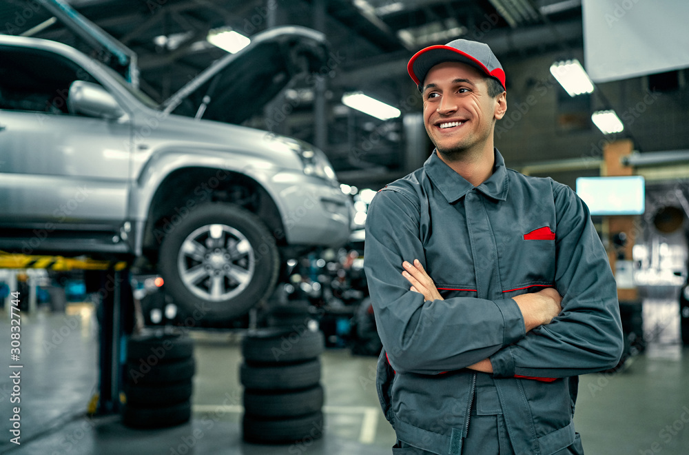 Poster handsome auto service mechanic in uniform is standing on the background of car with open hood, smili