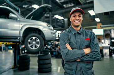 Handsome auto service mechanic in uniform is standing on the background of car with open hood, smiling and looking at camera. Car repair and maintenance.