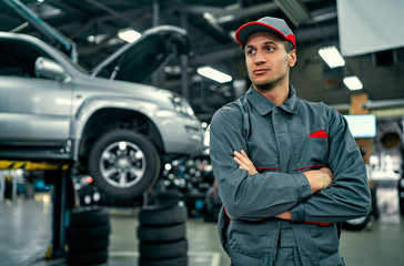 Handsome auto service mechanic in uniform is standing on the background of car with open hood and...