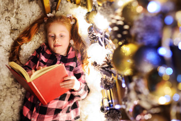 a little girl with red hair is lying under a Christmas tree, reading a book of fairy tales and...