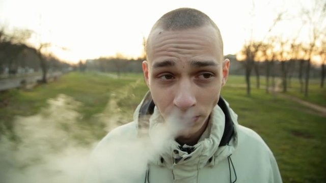 Funny wide angle closeup portrait of smoking e-cigarette young man in the park in the evening