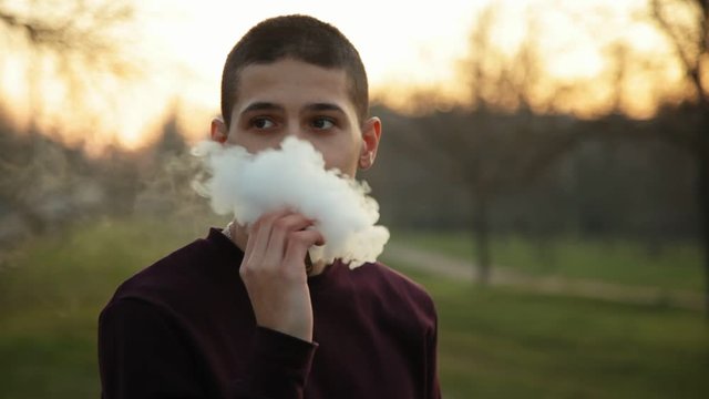 Pensive man smoking electronic cigarette and exhale clounds of vapor in the park at chilly late autumn evening, close up