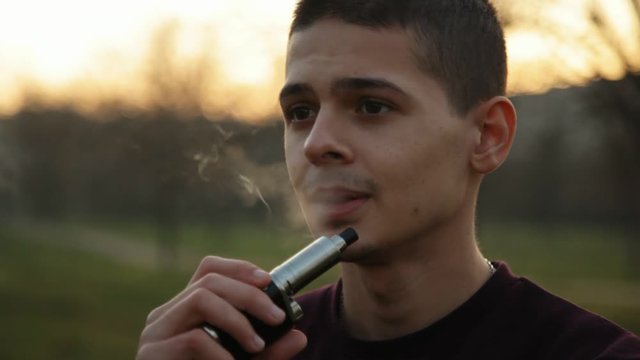 Relaxed caucasian young man with short hair vaping e-cigarette in the park in the evening, Close up