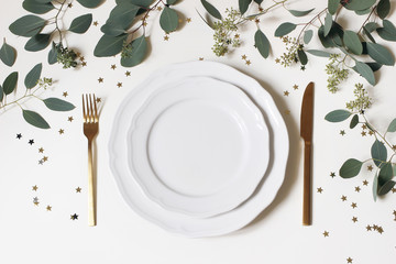 Christmas, New Year table place setting. Golden cutlery, porcelain plate, eucalyptus branches and golden confetti stars isolated on white background. Winter holidays background. Flat lay, top view.