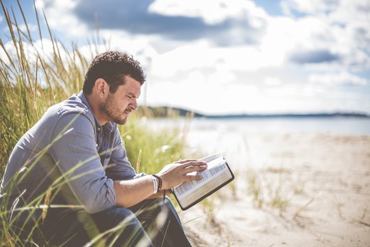 Shallow focus shot of a male sitting on the beach shore while reading the bible