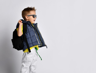 Attractive little boy in stylish warm clothes with a backpack on his shoulders, having fun on a light studio background.