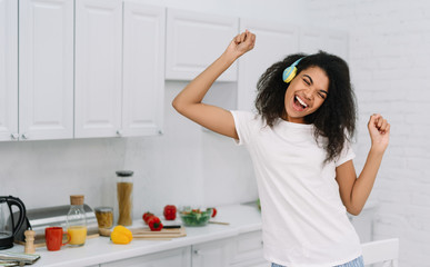 Happy emotional woman dancing at home, having fun. Portrait of beautiful African American girl listening music using wireless headphones