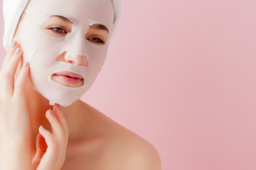 Beautiful young woman is applying a cosmetic tissue mask on a face on a pink background. Healthcare and beauty treatment and technology concept