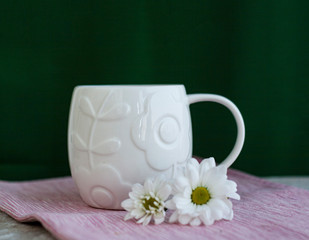 White cup stands on pink napkin on green background. Chrysanthemum flowers lie nearby. There is space for text on the background.