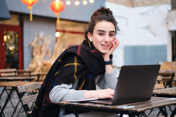 Pretty smiling student girl with scarf studying on laptop happily looking in camera in cafe on city street
