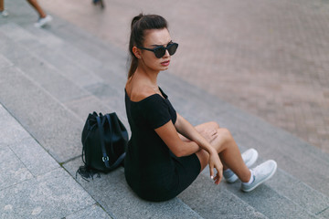 Fototapeta premium Lonely girl with frown face, sits on the stairs in the city, in a black dress with round sunglasses, looking after her shoulder Full length portrait.