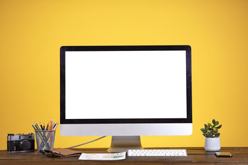 Workplace concept with blank white computer screen at wooden desk over pale yellow background, copy space