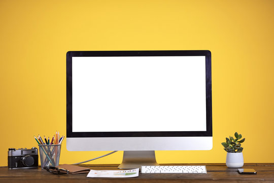 Workplace concept with blank white computer screen at wooden desk over pale yellow background, copy space
