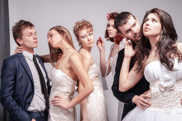Group of Five Loving Caucasian People Standing in Pairs of Two in Wedding Clothing. Against Gray Background