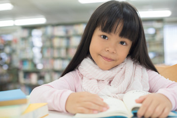 portrait of cute schoolgirl smiling while sitting with stack of books at table in library