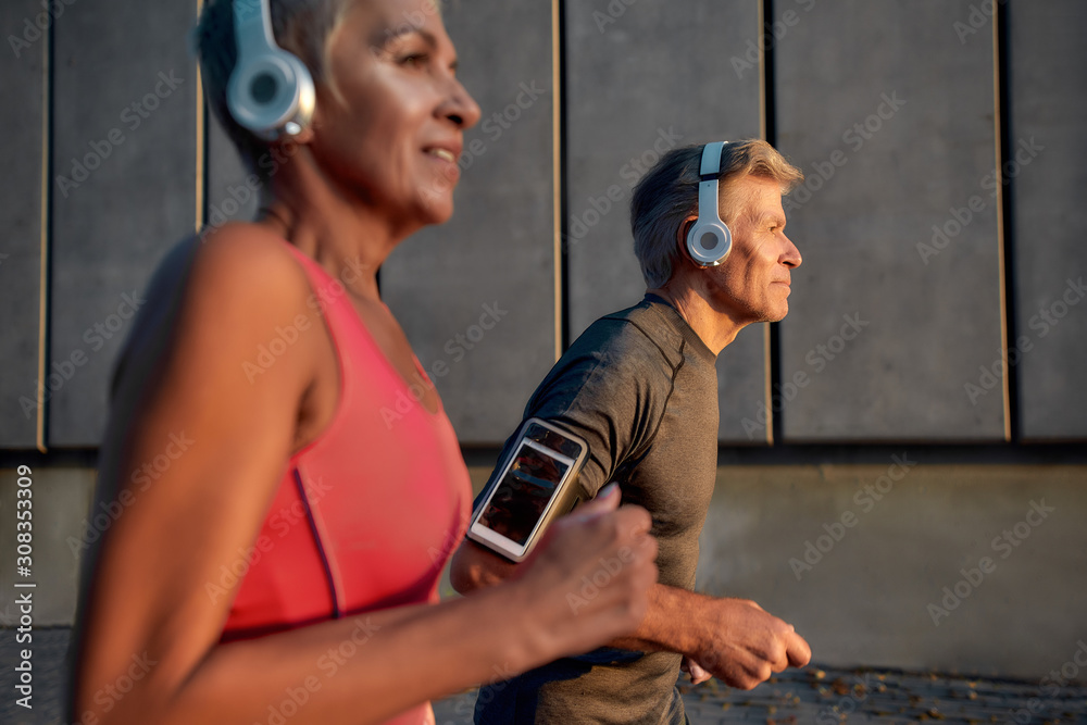 Wall mural Keep on moving. Sporty and happy middle-aged couple in headphones running together through the city street in the morning