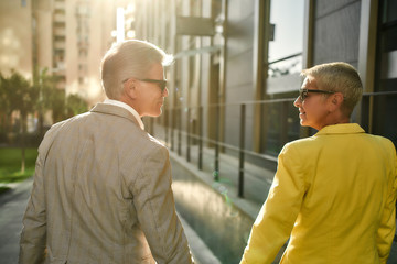 Obraz premium Great sunny day. Back view of stylish middle-aged couple in sunglasses holding hands and looking at each other while walking together through the city street