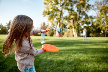 Making memories, breaking the distance. Little girl playing frisbee with her family in the park on a sunny day - obrazy, fototapety, plakaty