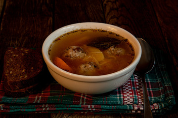 Homemade soup with meatballs in a white toe, on a kitchen checkered napkin with bread and grains and a teaspoon.