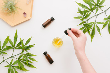 Woman's hand, glass bottles with CBD oil, THC tincture and hemp leaves on a white background. Flat lay, minimalism. Cosmetics CBD oil.