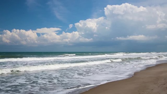 Ocean waves break on a sandy beach under a cloudy blue sky over a calm sea in this seamlessly looping video footage.