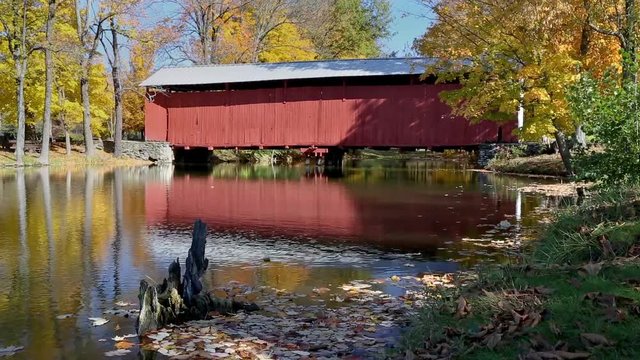 A reflection on a pond with ripples of a historic red covered bridge is surrounded by a beautiful autumn Indiana landscape in this looping video footage.