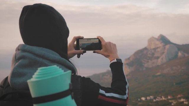 Girl on mountain top, female tourist taking photo of on cell smart phone. Young woman traveler on a background of mountains takes photos on a mobile phone.