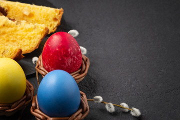 Painted Easter eggs in wicker coasters, pieces of Easter cake on a cutting board on a dark table - traditional Easter breakfast