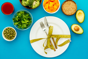 Diet concept. Fork and knife tied with measure tape on plate near vegetables on blue background top view