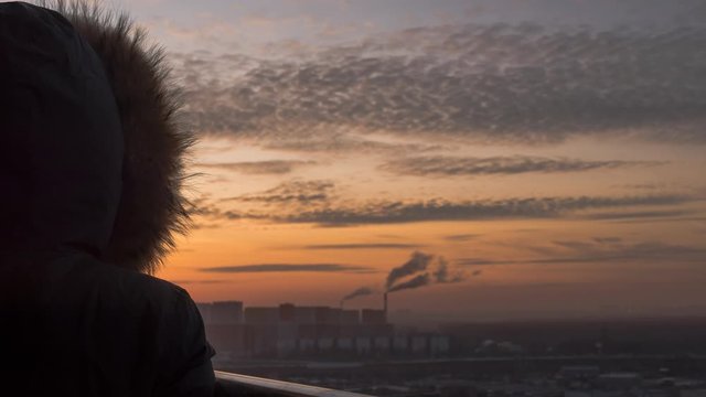 Time lapse of a woman in a winter mustard parka jacket with furry hood standing on the balcony of a northern city and looking on the power station and snowy town at sunset