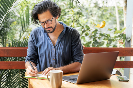 Handsome young curly latin man with glasses working on a laptop on the terrace of his house. Creative remote work, programmer, copywriter, writer