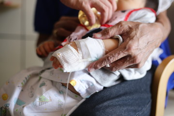 saline infusion. sick baby inward patient sitting on parent legs. baby in hospital being fed. sick kid in treatment process, in hospital and caring by many people.