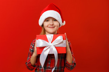 Cute happy child holds a box with a gift. Charming little girl in santa hat and dress on a red background. Christmas and New Year concept