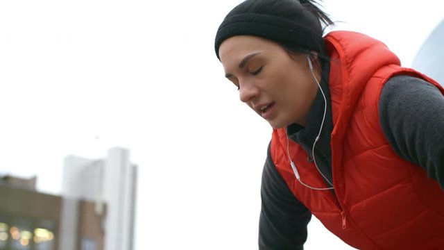 Medium shot of tired young female athlete in sportswear and earphones breathing heavily after outdoor workout