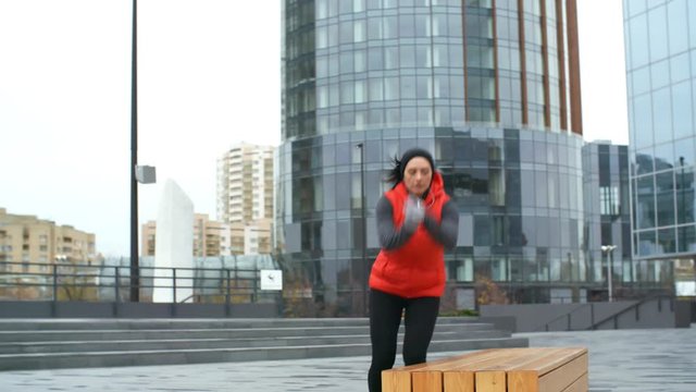 Front view of energetic young sportswoman doing jumping cardio workout with bench outdoors