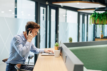 Fototapeta premium Serious young businessman talking on phone and reading e-mail on laptop screen when standing at bar counter