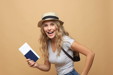 Beautiful blonde woman wearing summer clothes posing with passport with tickets over beige background.