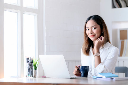 Portrait of young businesswoman smiling and looking at camera while sitting at office desk in modern office.