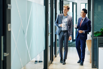 Multi-ethnic business people walking along the office corridor with cups of coffee and discussing...