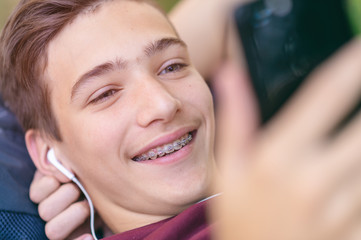 Happy teenage boy is using mobile phone, outdoors.  Close-up portrait of a smiling young man with smartphone, in park.  Cheerful teenager in casual clothes with cell phone in park. Soft focus