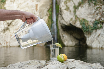 A hand pouring filtered water in the big glass with lemon on the nature background. Filter jug