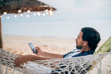 A man  lies in a hammock with a phone on the background of the ocean and sunset.