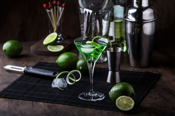 Green alcoholic cocktail martini glass with dry gin, vermouth, liquor, lime zest and ice, bar tools, dark background