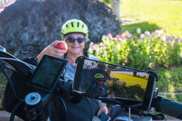 Healthy lifestyle. Attractive senior woman cyclist with yellow helmet have a break in a green park.  Moment of relaxation eating a red apple fruit.