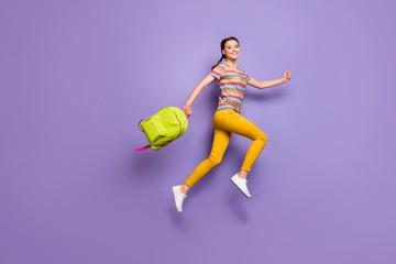 Fototapeta na wymiar Full size photo of crazy excited lady jumping high carry school green rucksack rushing home after lessons wear casual striped t-shirt yellow pants isolated purple color background