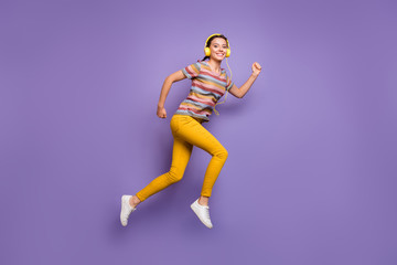Full body photo of pretty lady jump high listen cool earflaps songs run outdoors active way of life wear casual striped t-shirt yellow trousers isolated purple color background