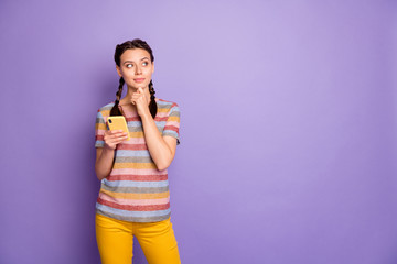 Portrait of minded interested girl teen use her smartphone think thoughts blogging touch chin look copyspace wear yellow pants outfit isolated over purple color background