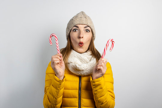 Young woman with a surprised face in a hat and a down jacket holds a candy caramel cane on a light background. Heart shape. Concept winter, christmas, winter holidays