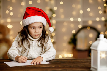 Happy little child girl in santa hat writing wish list or letter to Santa Claus on background with lights. Merry Christmas